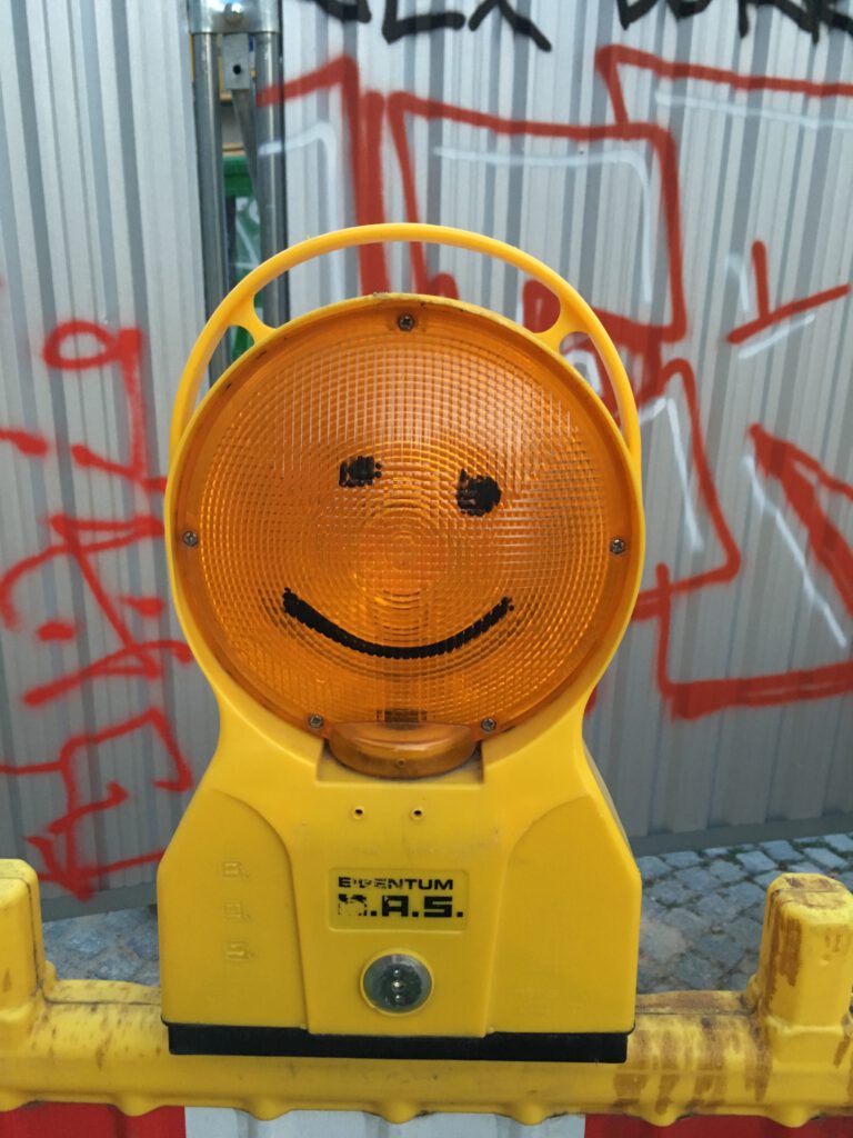 Orange Baustellenlampe, auf die mit schwarzem Stift ein einfaches, lächelndes Gesicht gemalt ist.